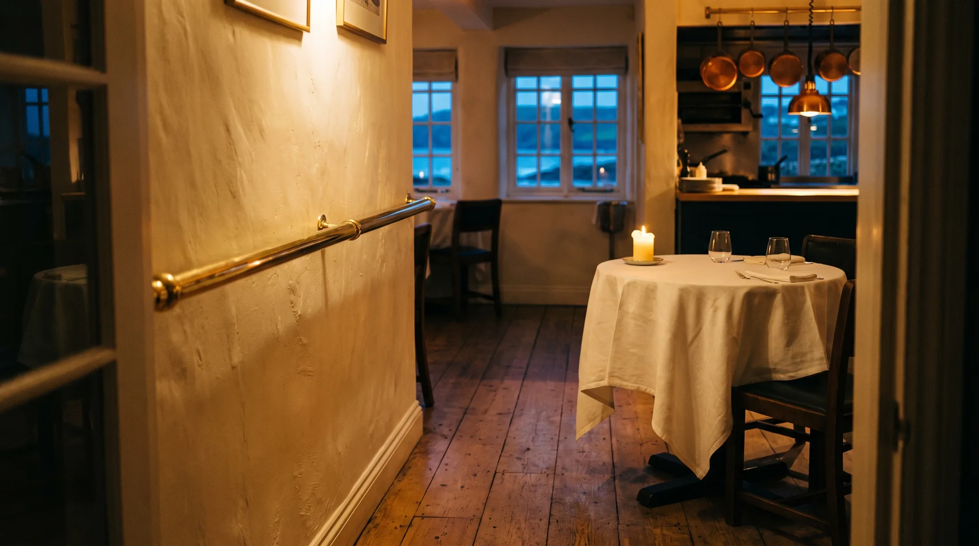 The dining room seen from just inside the door: a brass rail along the cream wall and a candlelit table for two.