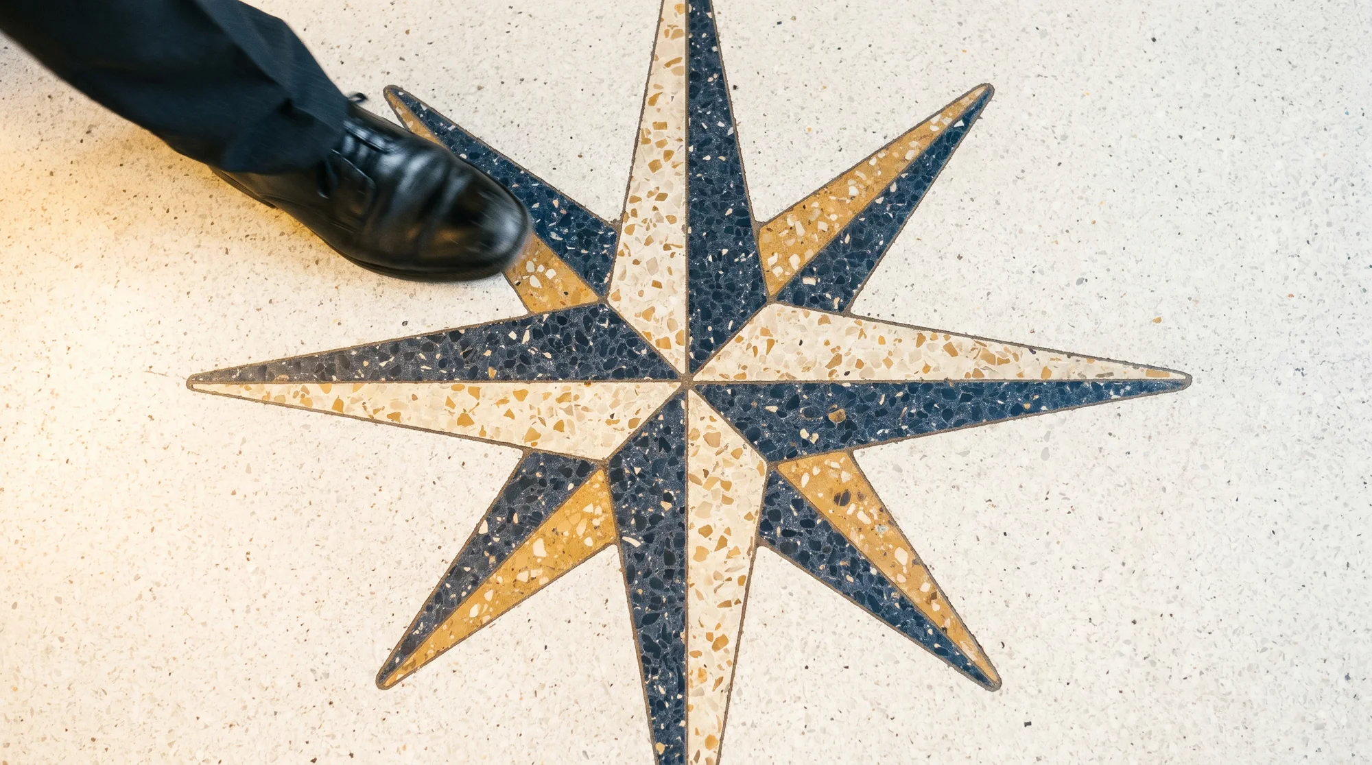 A compass rose inlaid in worn terrazzo, a waiter's shoe passing through the top of the frame.